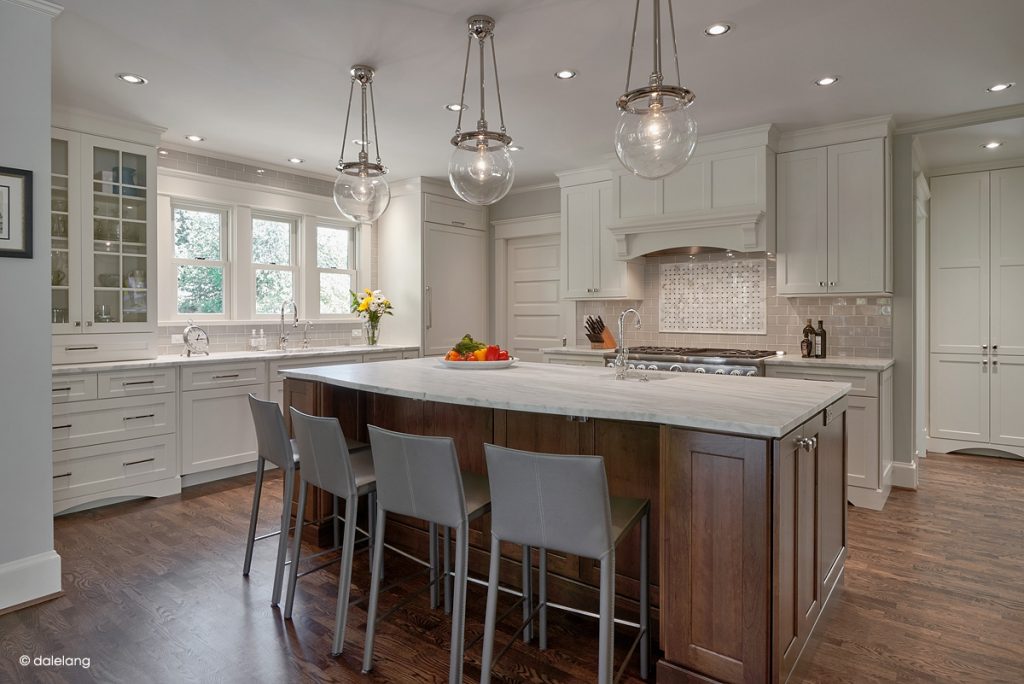 Modern kitchen with white cabinets, large island with marble countertop, four gray chairs, pendant lights, stainless appliances, and wooden floors. A bowl of fruit sits on the island; windows provide natural light.
