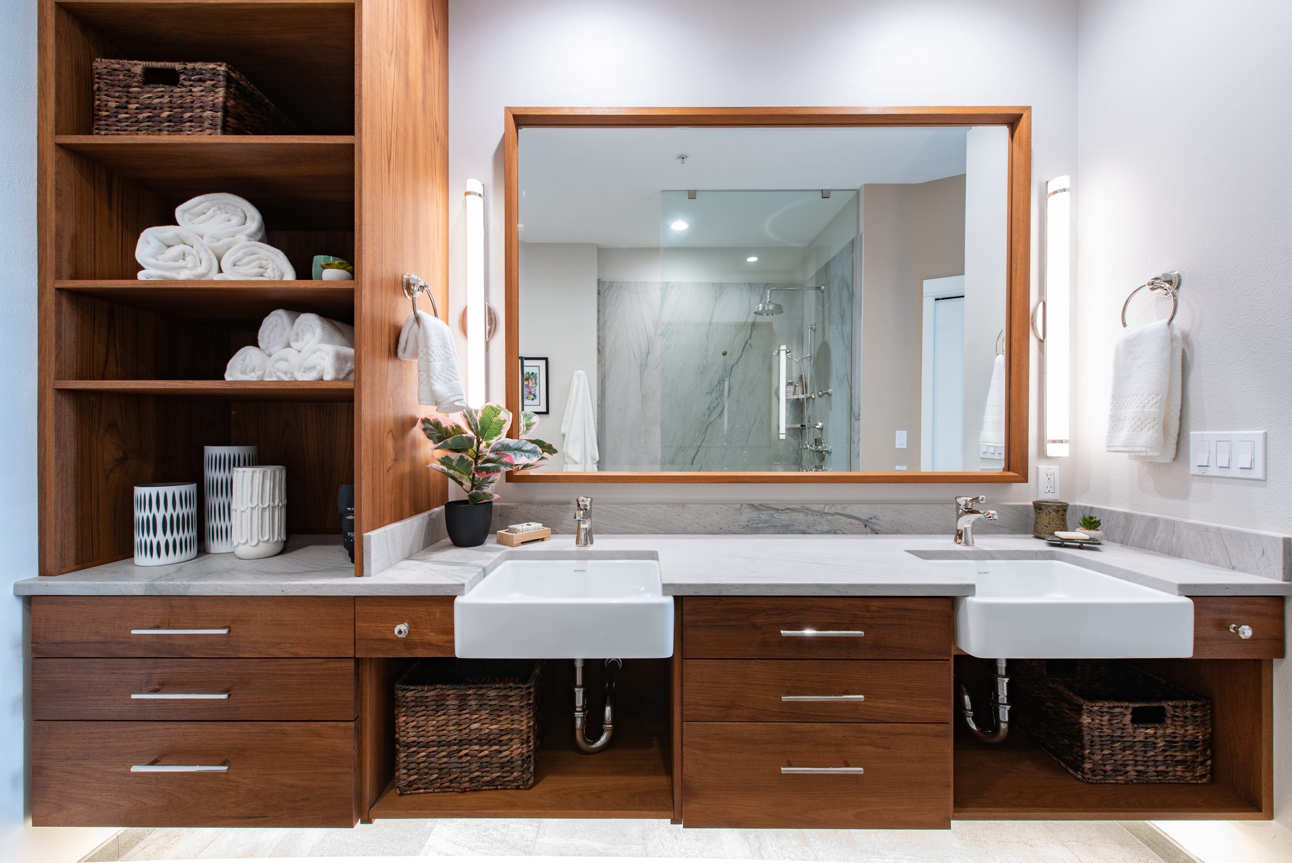 Modern bathroom with double sinks, wooden cabinets, open shelves holding towels and baskets, large mirror, wall lights, and a glass-enclosed shower reflected in the mirror. A small plant sits between the sinks.