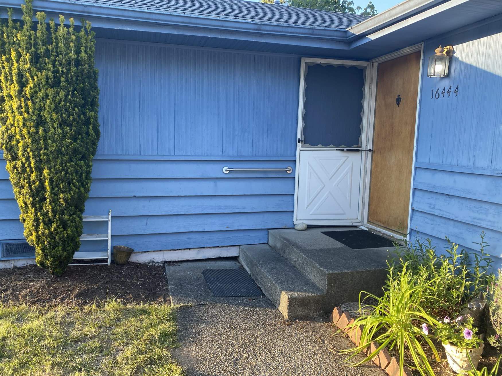 A blue house with a wooden door, a white half screen door, a small front porch with steps, a handrail, a potted plant, a bush, a small white ladder, and the number 16444 visible on the wall.