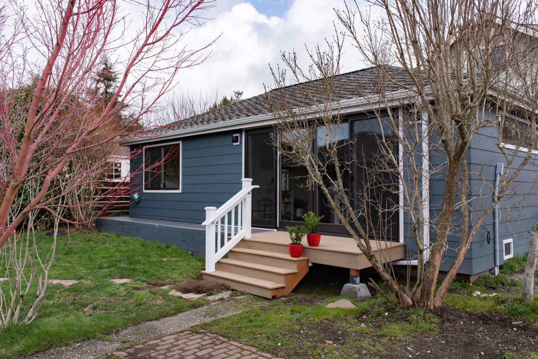 backyard-cottage-potter-construction-seattle-scaled A small blue house with white trim, a wooden porch with white railings, red potted plants, and a grassy yard surrounded by leafless trees and shrubs on a cloudy day.