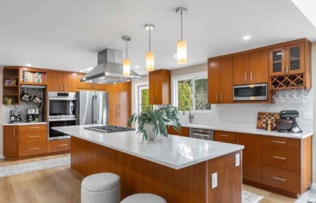 Modern Black Walnut Kitchen with wooden cabinets, white countertops, stainless steel appliances, island with stools, pendant lights, plant centerpiece, large windows, and built-in shelves displaying kitchen items.