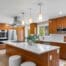 Modern Black Walnut Kitchen with wooden cabinets, white countertops, stainless steel appliances, island with stools, pendant lights, plant centerpiece, large windows, and built-in shelves displaying kitchen items.