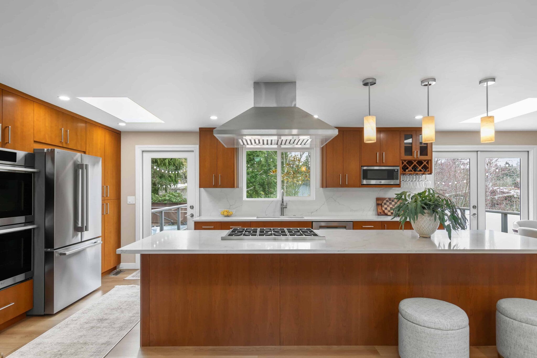 11114-34th-Pl-SW-Seattle-WA_0002-scaled Modern Black Walnut Kitchen with wood cabinets, stainless steel appliances, large island with white countertop, gas stove, hanging lights, and window overlooking greenery. Two round stools sit at the island.