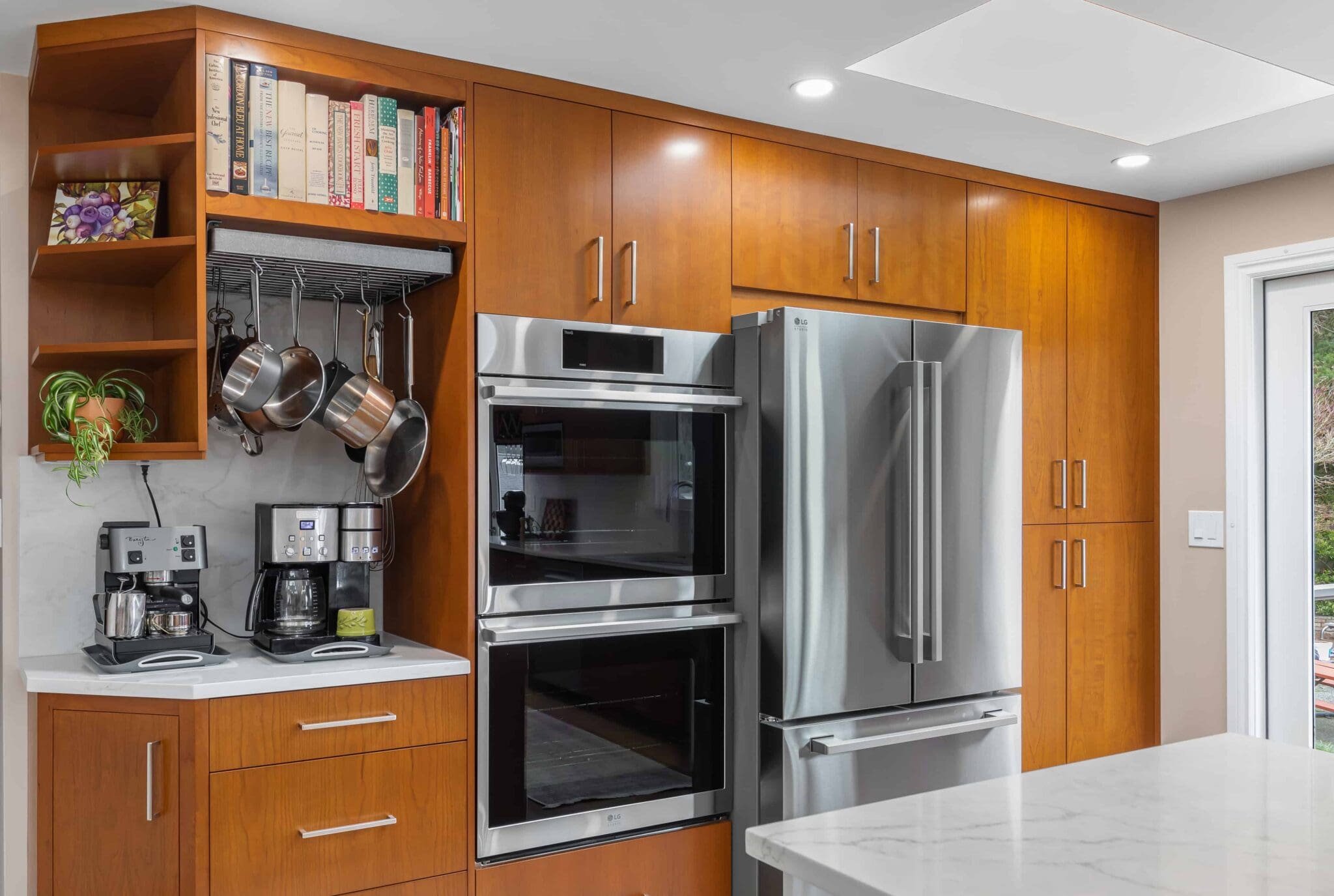 11114-34th-Pl-SW-Seattle-WA_0004-scaled Modern Black Walnut Kitchen with wood cabinets, stainless steel double oven, French door refrigerator, coffee makers, hanging pots, potted plants, and cookbooks on white counters under bright recessed lights.
