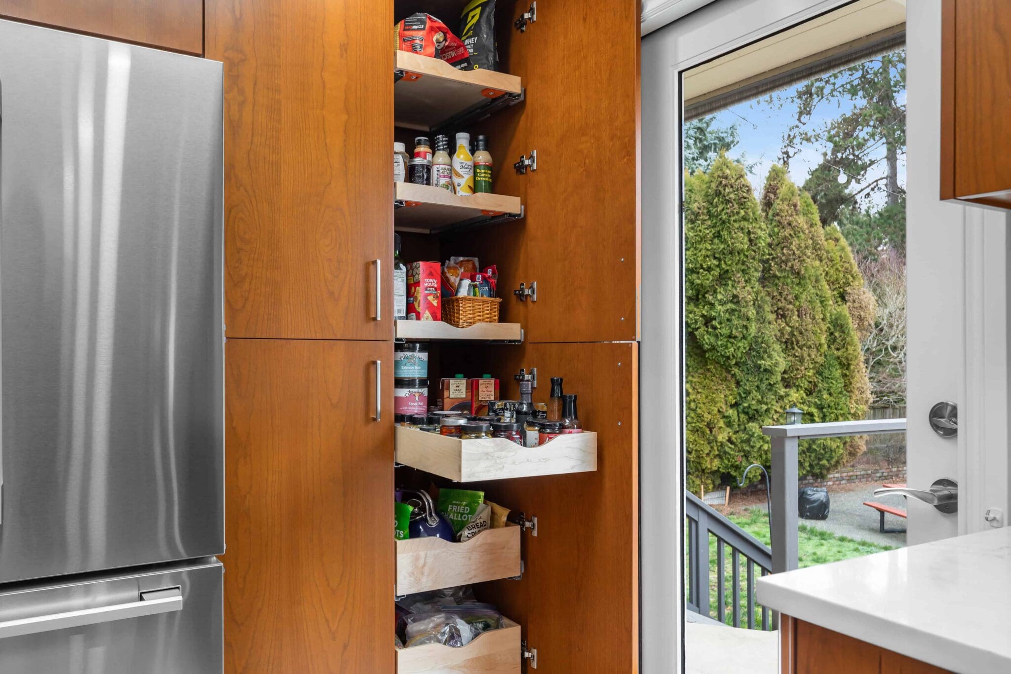 11114-34th-Pl-SW-Seattle-WA_0005-scaled A Black Walnut Kitchen pantry with wooden pull-out shelves is open, displaying various food items. Beside it stands a stainless steel refrigerator and a glass door leading to a green backyard.