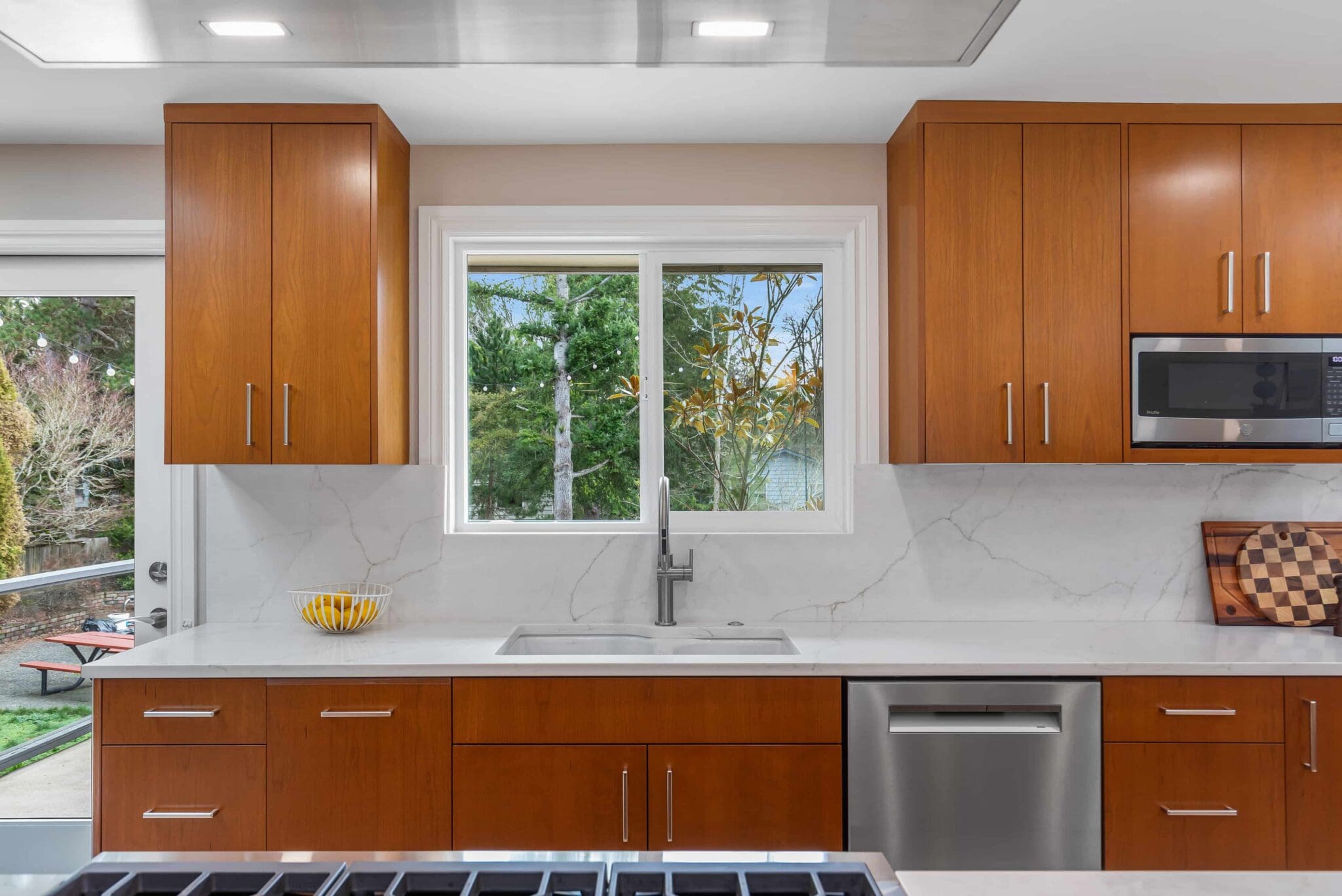 11114-34th-Pl-SW-Seattle-WA_0007-scaled Modern Black Walnut Kitchen with wood cabinets, white countertops, stainless steel dishwasher, built-in microwave, sink under a window, bowl of lemons, sliding glass door, and cutting board.
