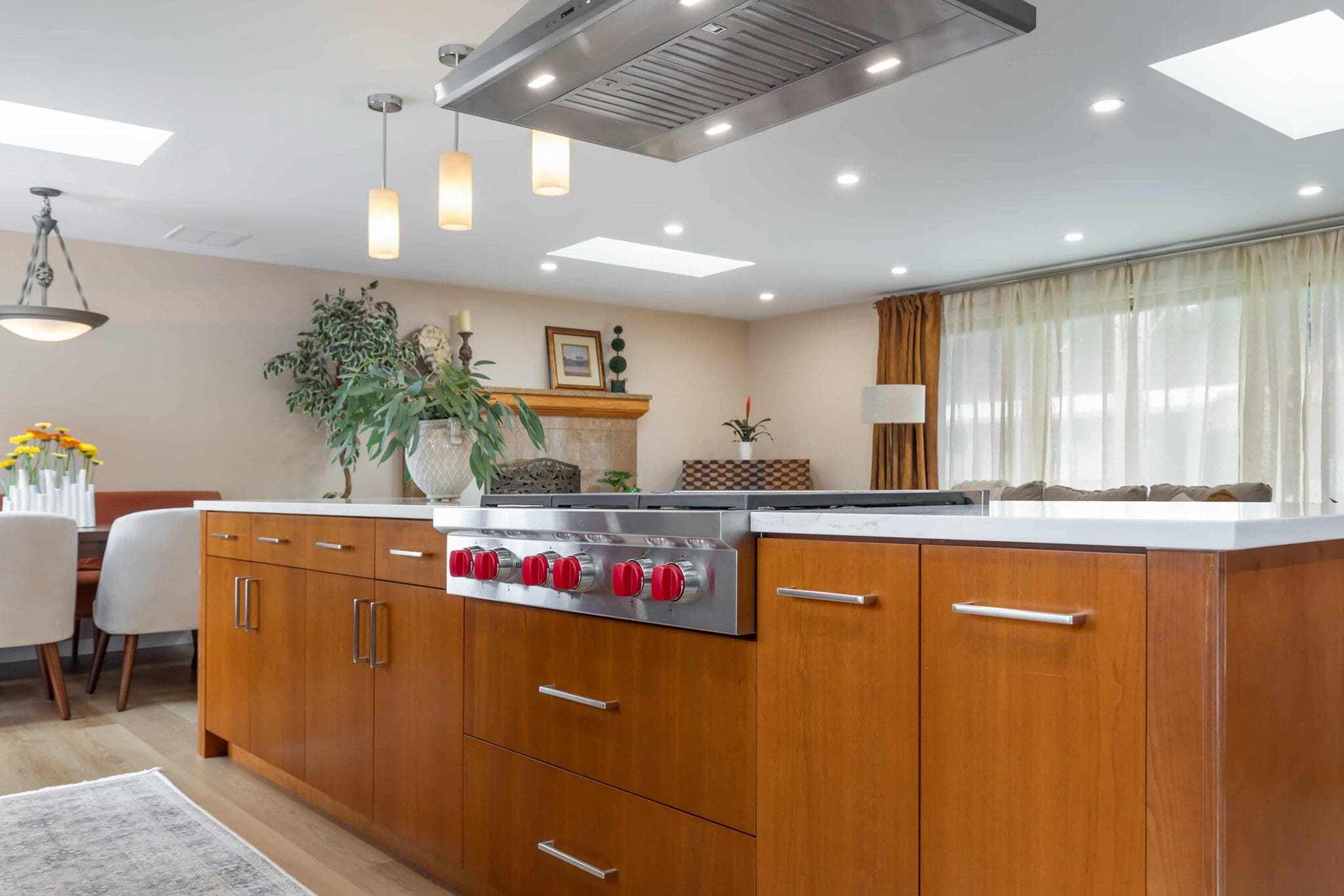 11114-34th-Pl-SW-Seattle-WA_0011-scaled Modern Black Walnut Kitchen with wooden cabinets, a stovetop with red knobs, hanging lights, indoor plants, and large windows with sheer curtains. Dining area with a table and chairs is visible in the background.