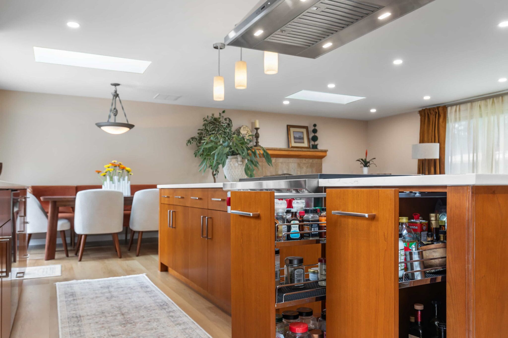 11114-34th-Pl-SW-Seattle-WA_0012-scaled Modern Black Walnut Kitchen with wooden cabinets, open spice rack drawer, white countertop, pendant lights, and a dining area featuring a table, white chairs, and a vase of yellow flowers in the background.