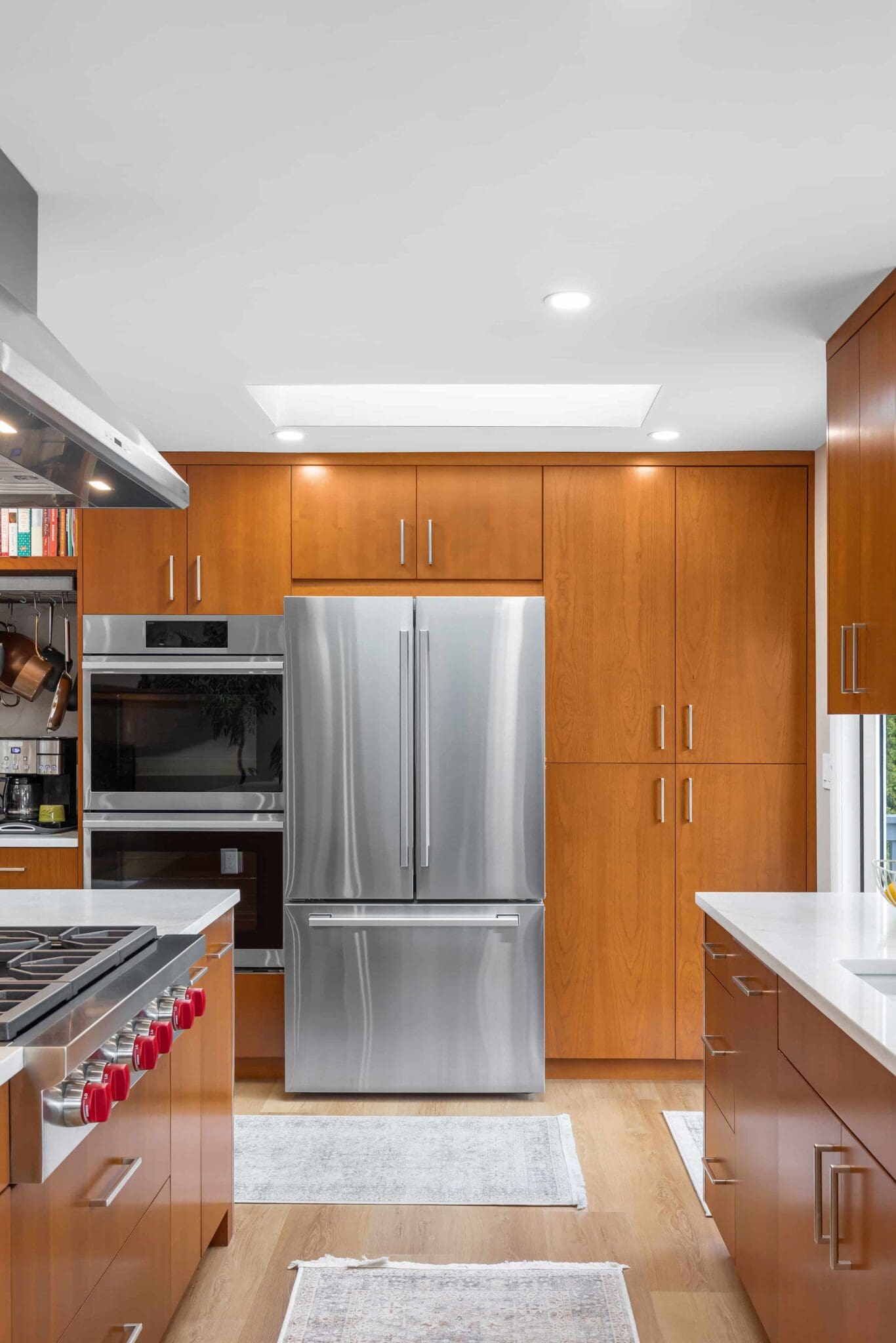 11114-34th-Pl-SW-Seattle-WA_0016-scaled Modern black walnut kitchen with wooden cabinets, stainless steel French door refrigerator, built-in oven, gas stove with red knobs, white countertops, and light wood flooring with white rugs.