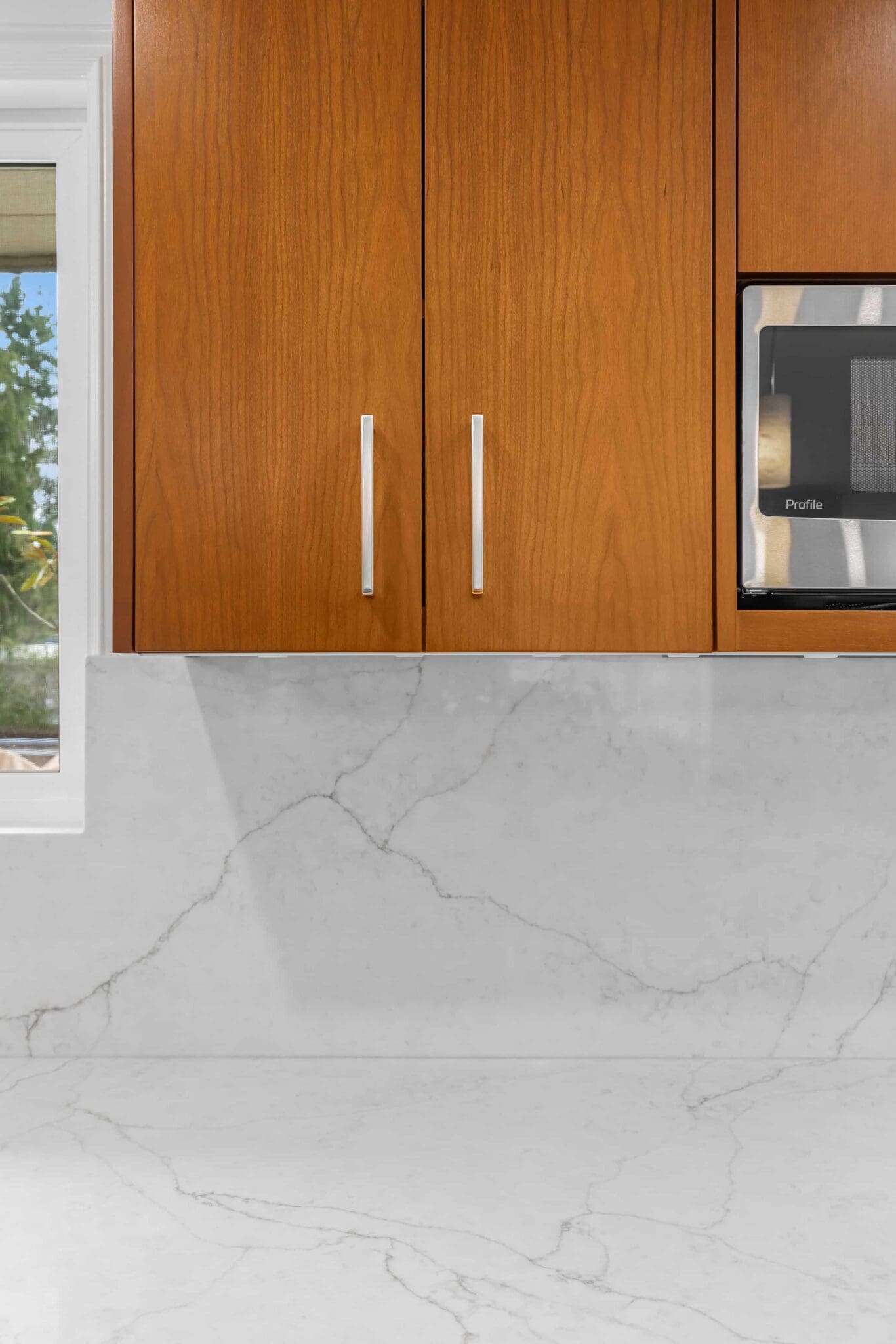 11114-34th-Pl-SW-Seattle-WA_0017-scaled Light gray marble countertop and backsplash with subtle veining, Black Walnut Kitchen upper cabinets with silver handles, window on the left, and a microwave partially visible on the right.
