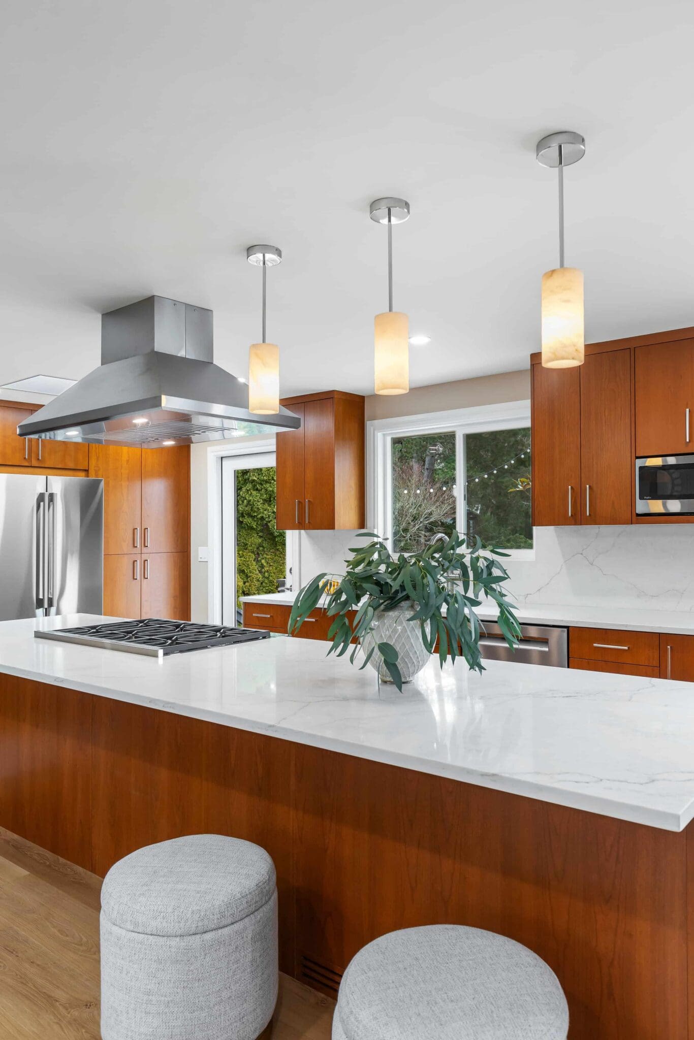 11114-34th-Pl-SW-Seattle-WA_0018-scaled Modern Black Walnut Kitchen with wood cabinets, stainless steel appliances, white marble countertops, three pendant lights, two round stools, and a green plant on the island beneath a large vent hood.