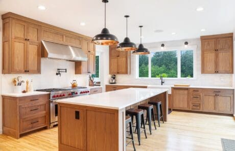 Modern cherry kitchen with wood cabinets, white countertops, a large island with four stools, black pendant lights, stainless steel appliances, and a large window overlooking greenery.