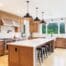 Modern cherry kitchen with wood cabinets, white countertops, a large island with four stools, black pendant lights, stainless steel appliances, and a large window overlooking greenery.