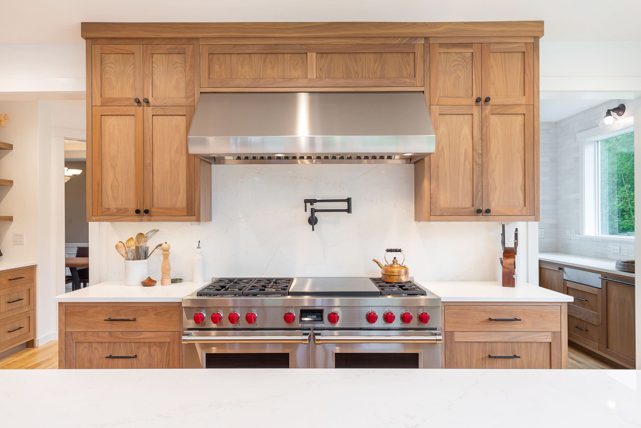 15313-241st-St-SE-Snohomish-WA_0005 Modern cherry kitchen with wood cabinets, a stainless steel range hood, red-knobbed stove, marble backsplash, pot filler, and countertop utensils. Bright and spacious with natural light from a window.