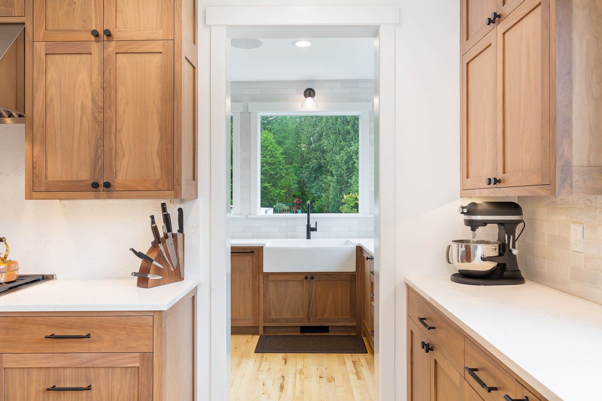 15313-241st-St-SE-Snohomish-WA_0010 A modern Cherry Kitchen with wooden cabinets, white countertops, a knife block, stand mixer, and a view into a bright room with a farmhouse sink and large windows showing green trees outside.