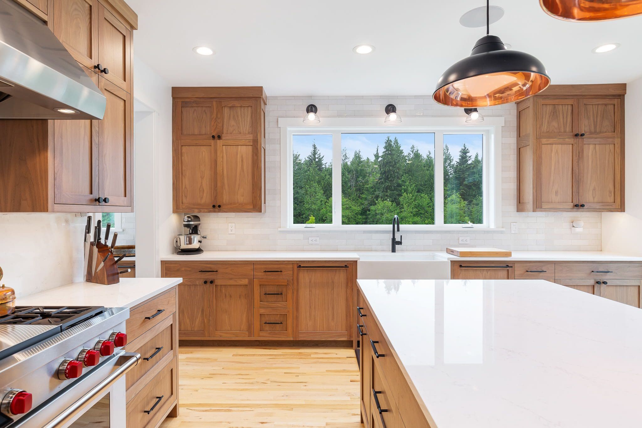 15313-241st-St-SE-Snohomish-WA_0012 Modern cherry kitchen with wooden cabinets, white countertops, a gas stove with red knobs, a large window showing trees outside, and black pendant light fixtures over a spacious island.