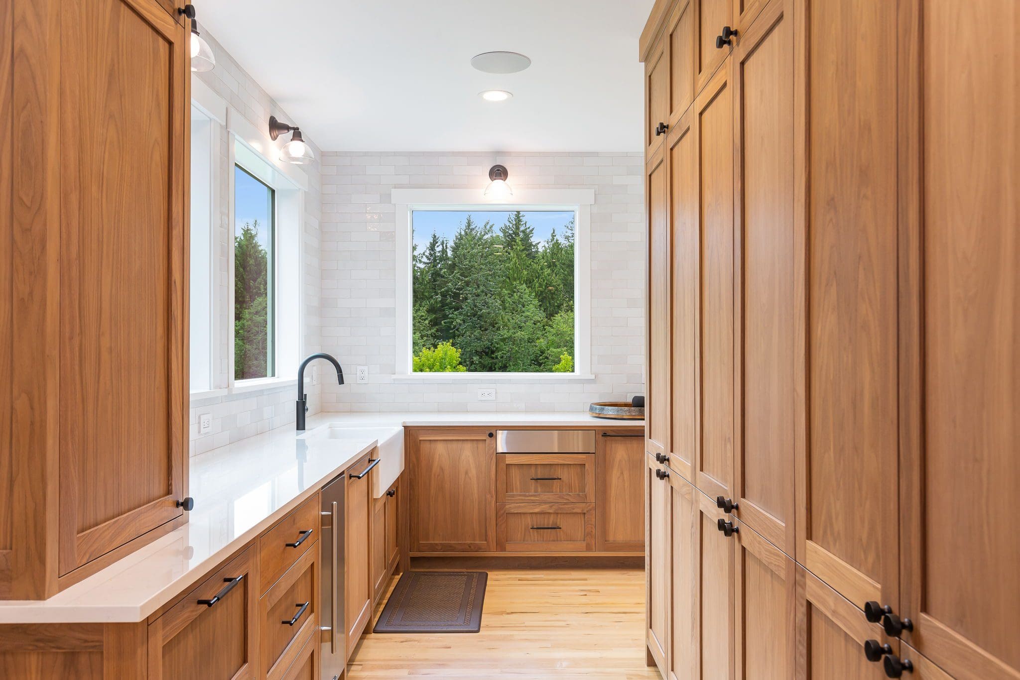 15313-241st-St-SE-Snohomish-WA_0017 Modern kitchen with light wood cabinets, white countertops, a black faucet, and large windows showing green trees outside. The bright cherry kitchen vibe is enhanced by a white subway tile backsplash.