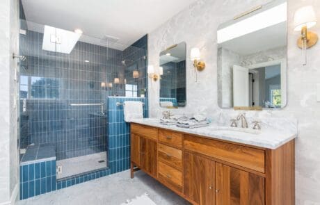 Modern bathroom with a wood double vanity, marble countertop, dual mirrors, wall sconces, and a large glass shower with blue tile. Natural light enters through a skylight above the shower.