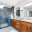 Modern bathroom with a wood double vanity, marble countertop, dual mirrors, wall sconces, and a large glass shower with blue tile. Natural light enters through a skylight above the shower.
