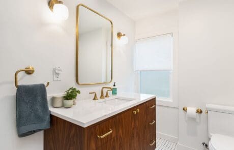 Modern bathroom with wood vanity, gold fixtures, rectangular mirror, two wall lights, a towel ring with a gray towel, and a small plant on the counter. White walls and window with shade, toilet and TP holder on the right.
