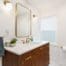 Modern bathroom with wood vanity, gold fixtures, rectangular mirror, two wall lights, a towel ring with a gray towel, and a small plant on the counter. White walls and window with shade, toilet and TP holder on the right.
