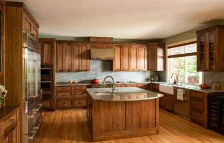 Spacious kitchen with wooden cabinets, a central island with a marble countertop, stainless steel appliances, hardwood floors, and large windows letting in natural light.