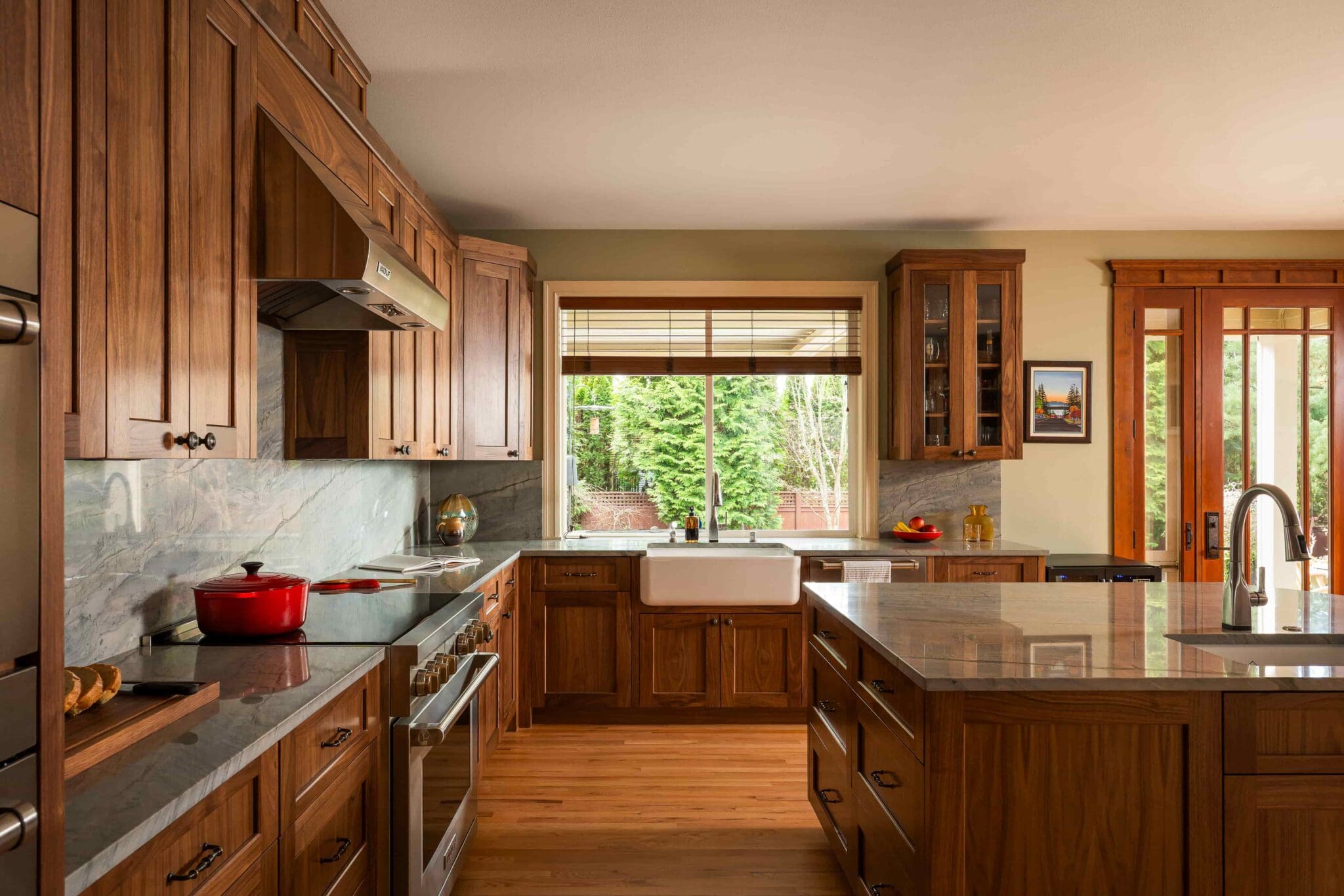 HAAS-Seattle-Kitchen-Remodel-10-2-scaled Modern kitchen with wooden cabinets, marble countertops, a farmhouse sink below a large window, stainless steel appliances, and a kitchen island, all warmly lit by natural light from the windows and glass door.