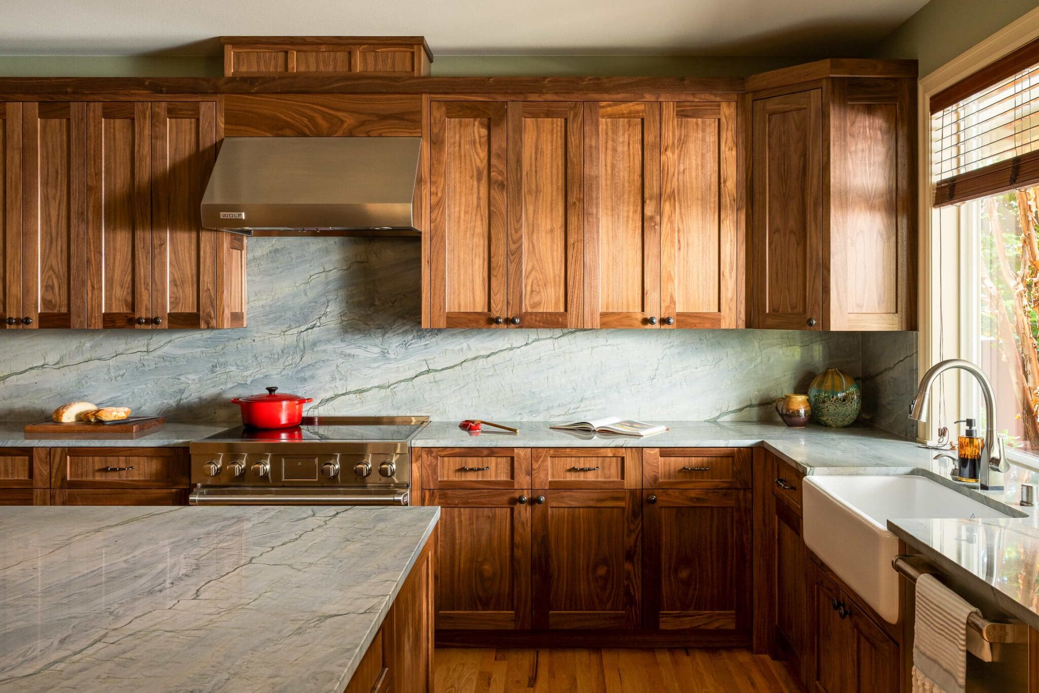 HAAS-Seattle-Kitchen-Remodel-3-2-scaled A kitchen with wooden cabinets, marble countertops and backsplash, a farmhouse sink, a stove with a red pot, and sunlight coming through a window with blinds.