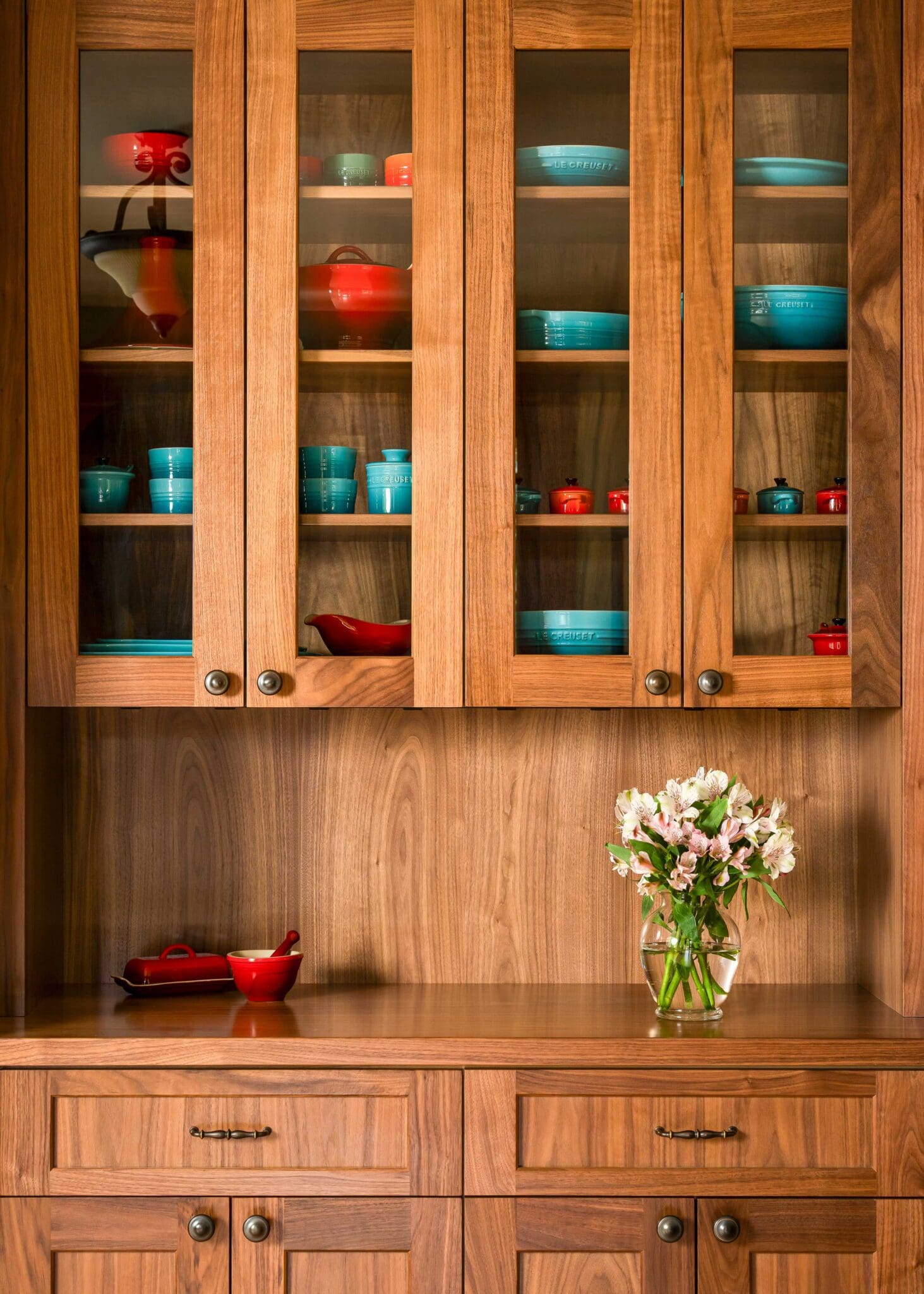 HAAS-Seattle-Kitchen-Remodel-5-2-scaled Wooden kitchen cabinet with glass doors displays turquoise blue and red dishes. Below, a wooden countertop holds a vase with pink and white flowers and a red dish. Drawer and cabinet doors are closed.