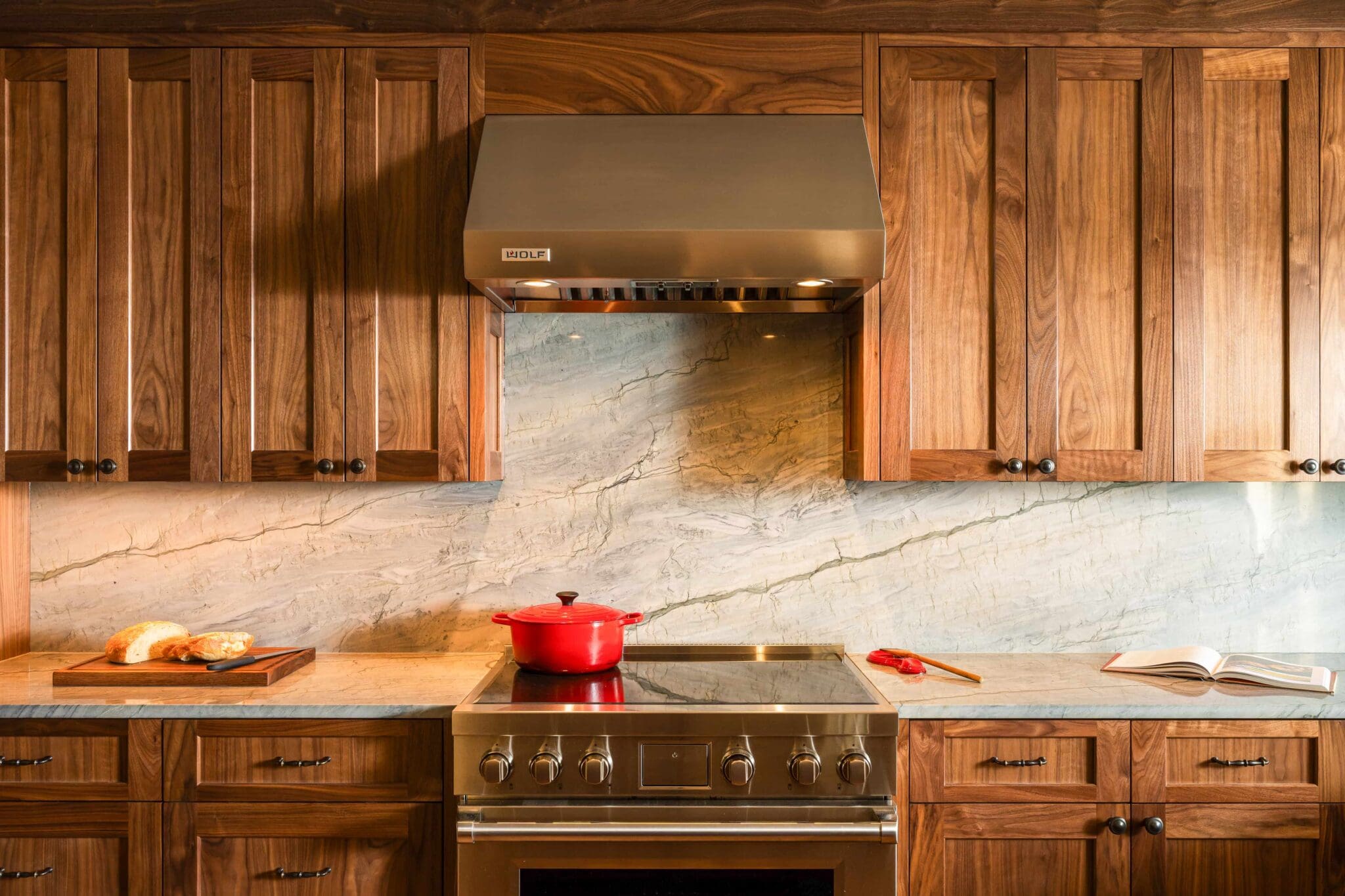 HAAS-Seattle-Kitchen-Remodel-7-2-scaled A kitchen with wooden cabinets, marble backsplash, stainless steel stove, and range hood. A red pot is on the stove, bread on a cutting board, and an open book with a red spatula nearby.