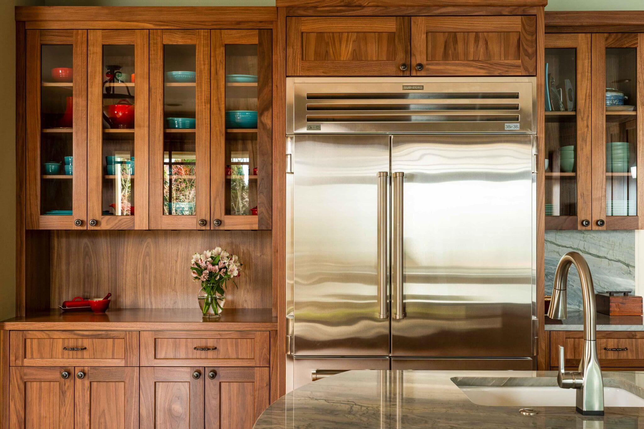 HAAS-Seattle-Kitchen-Remodel-8-2-scaled Modern kitchen with wood cabinets, glass cabinet doors displaying colorful dishes, a large stainless steel double-door refrigerator, marble countertop, and a sink with a curved faucet; a vase of flowers sits on the counter.