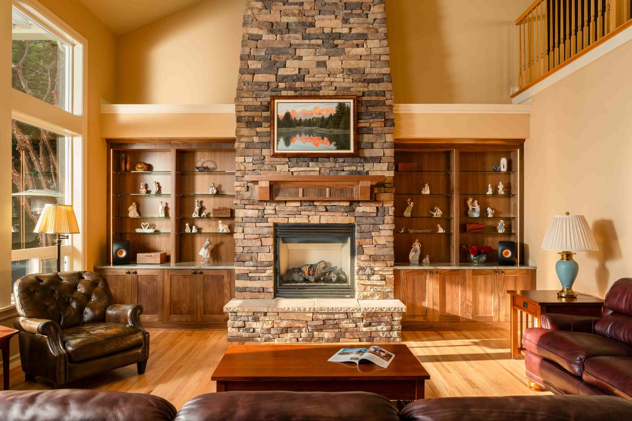 HAAS-Seattle-Living-Room-Fireplace-Remodel-14-1-scaled A cozy living room with a stone fireplace, wooden shelves holding decor, leather chairs, a wooden coffee table with an open magazine, and warm lighting from table lamps. A landscape painting hangs above the fireplace.