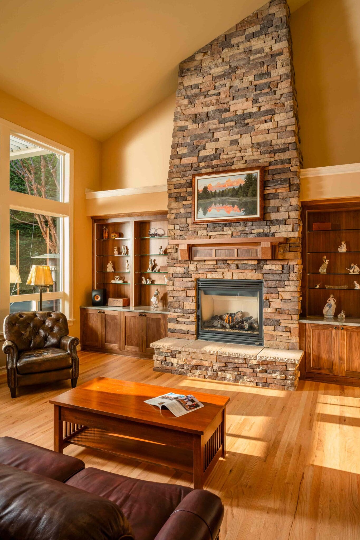HAAS-Seattle-Living-Room-Fireplace-Remodel-15-2-scaled A cozy living room with a tall stone fireplace, wood mantel, framed art, built-in shelves, and leather chairs. Sunlight streams through large windows onto a wooden coffee table with an open magazine.