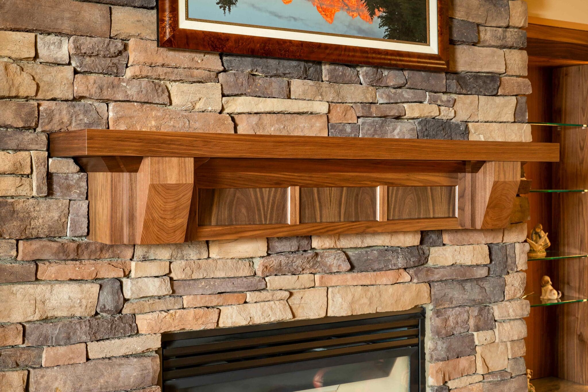 HAAS-Seattle-Living-Room-Fireplace-Remodel-17-2-scaled A wooden mantel is mounted on a stone fireplace with a glass-fronted firebox below. The mantel is rectangular with square accents, and a framed picture hangs above it on the stone wall. Shelves are visible on the right.