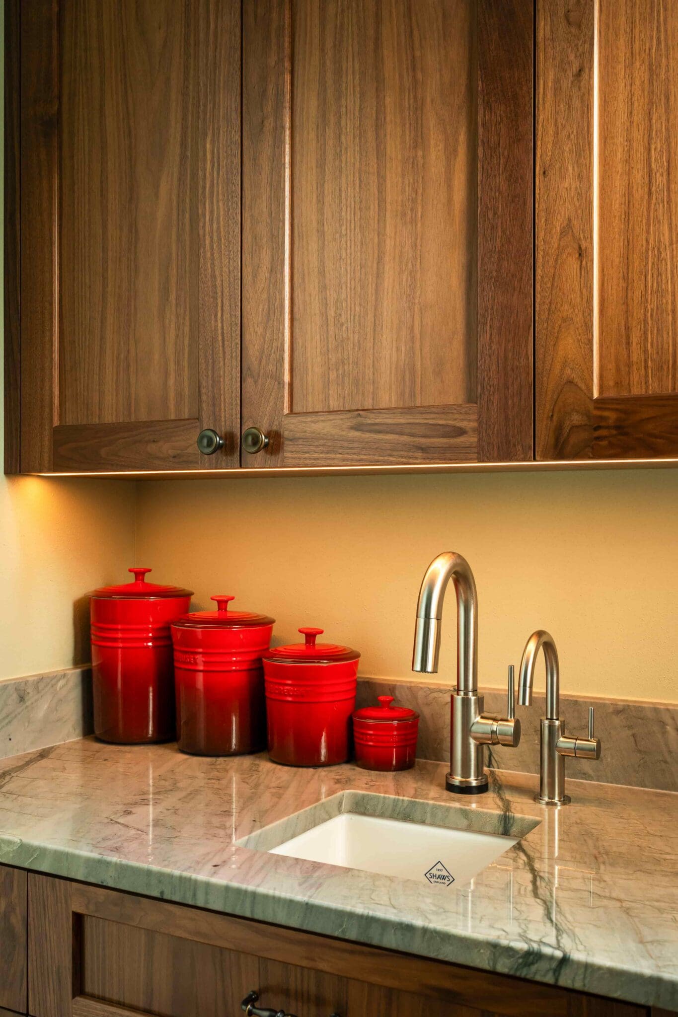 HAAS-Seattle-Pantry-Remodel-12-1-1-scaled A kitchen sink with stainless steel faucets, marble countertop, wooden cabinets, and four red storage canisters arranged next to the sink under warm lighting.